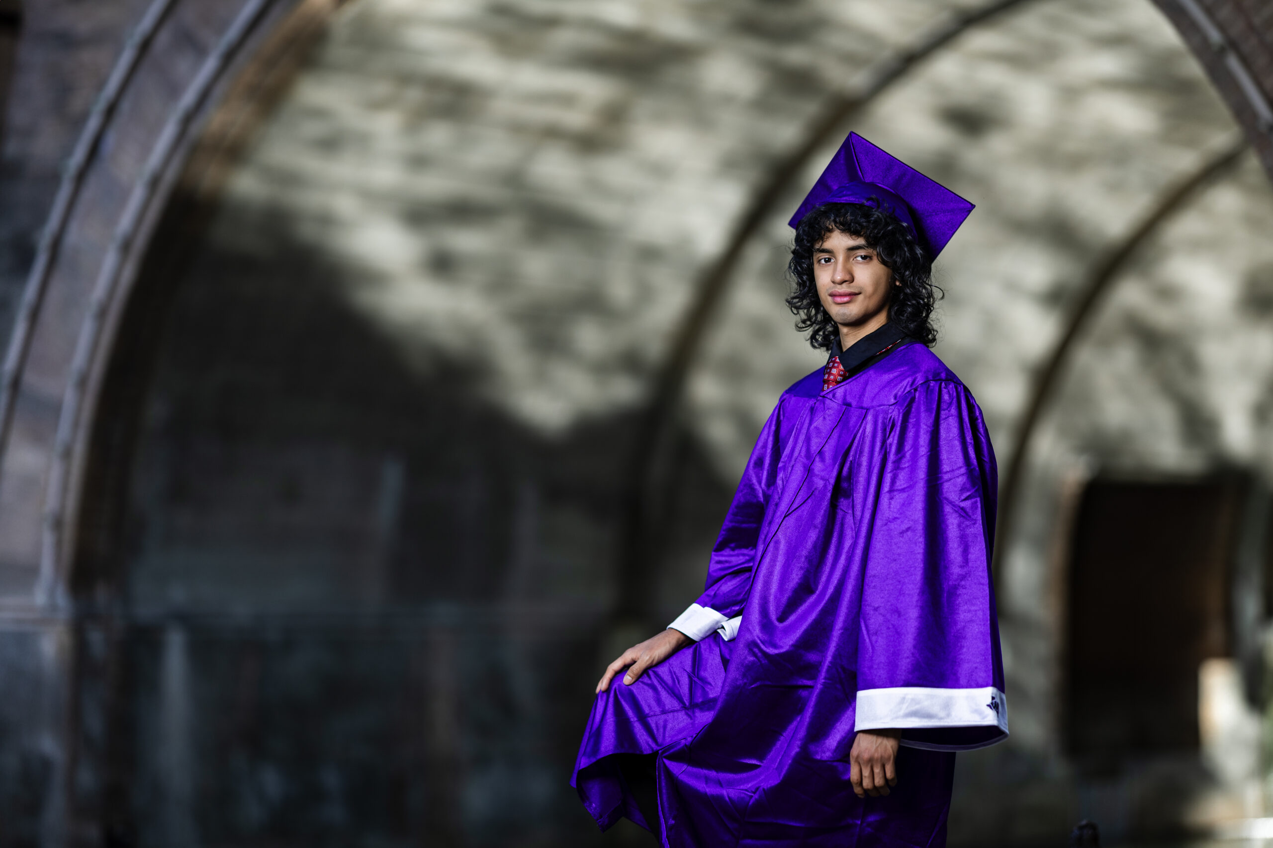 Graduate in purple cap and gown posing by stone arches – Cabrera Photography Graduation